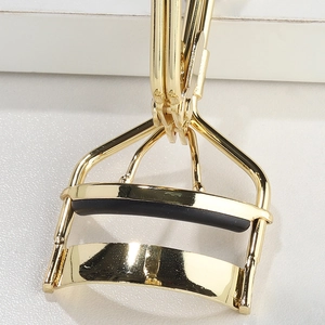 Close-up of golden stainless steel eyelash curler with black rubber pad on white surface
