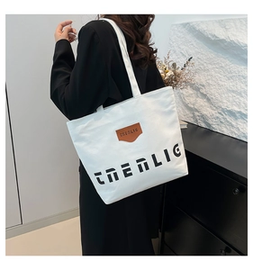 White canvas tote bag with black letter print and brown leather tag carried by model