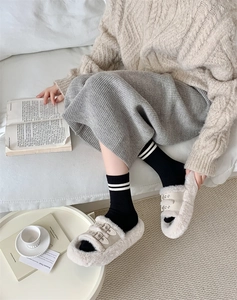 Close-up of beige plush slippers with metal buckle worn by a model in a cozy home setting