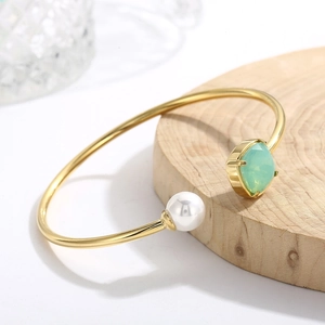 Dettaglio del bracciale dorato a C con perla bianca e gemma verde su vassoio di legno