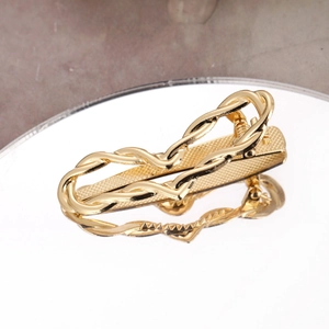 Elegant gold metal tie clip with textured grid pattern and twisted design on a mirror stand