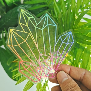 Hand holding a rainbow holographic geometric crystal suncatcher sticker against green leaves