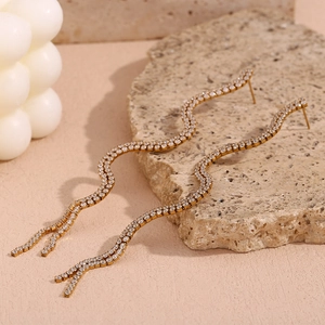 Macro detail of gold S-shaped wave earrings with white zircons on beige marble
