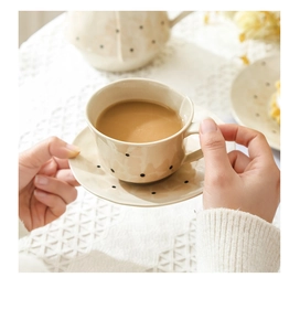 Gros plan d'une tasse à café en céramique beige à pois noirs tenue à la main sur nappe