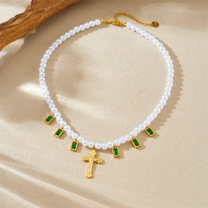 Vintage pearl necklace with green square gems and a gold cross pendant on beige surface