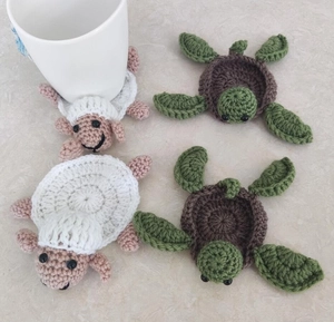 Close-up of handmade crochet turtle and sheep coasters with white mug on marble top