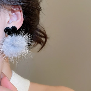 Dettaglio di orecchini a cuore con pompon in peluche grigio indossati su una modella