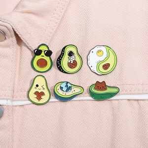 Set of six creative avocado enamel pins displayed on pink denim fabric background