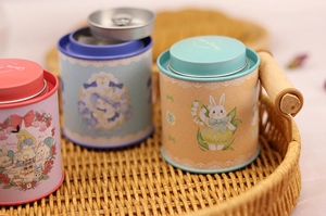 Three colorful tinplate tea canisters with cute illustrations on a woven tray