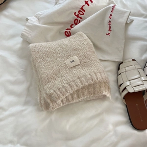 Detail of a beige knit wool scarf with a small logo tag on a white fabric background
