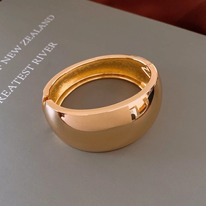 Close-up of a wide polished gold bangle with a hinged closure on a grey book background