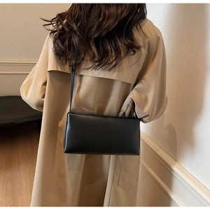 Detail of a minimalist black leather crossbody bag worn by a model in a beige trench coat