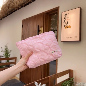 Pink pleated textured cosmetic bag with brand label shown in a lifestyle outdoor setting