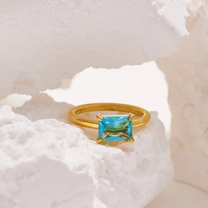 Close-up of an 18K gold plated ring with a teal emerald-cut stone on a white stone base