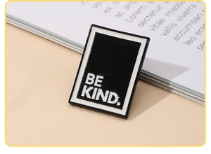 Close-up of rectangular black enamel pin with white Be Kind text on a magazine background