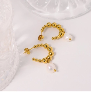 Detail view of gold beaded C-shape earrings with hanging white pearls on a glass prop