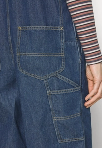 Detail view of blue denim overalls showing cargo pocket and yellow double stitching