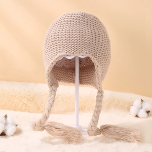 Detail of beige knit baby hat with earflaps, soft textures, and braided tassels on plush rug