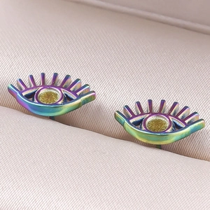 Macro shot of rainbow stainless steel evil eye earrings on a beige quilted surface