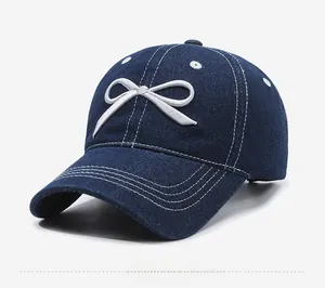 Detail view of washed blue denim baseball cap featuring a white embroidered bow and stitching