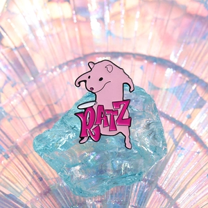 Pink cartoon rat enamel pin with RATZ text displayed on a crystal ice block base