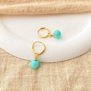 Close-up of sky blue round pearls on 18K gold plated hoops over a beige linen background