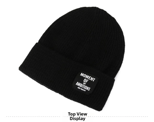 Top view of a black knitted beanie featuring a square patch with white artistic lettering