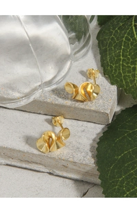Detailed view of gold ribbon stud earrings on a grey stone base with green leaves