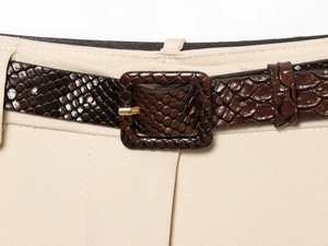 Détail d'une ceinture marron foncé aspect serpent avec boucle dorée sur un pantalon beige