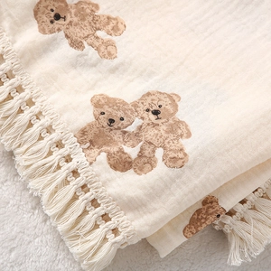Detailed close-up of beige cotton muslin baby blanket with bear print and fringe trim
