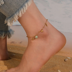 Close-up of a gold chain anklet featuring fish charms and a green bead on the beach