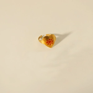 Detailed close-up of golden heart-shaped open ring with red sun pattern on beige background
