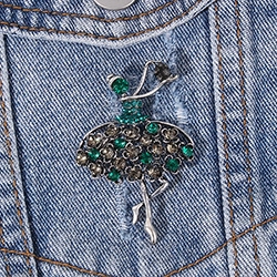 Close-up of a silver ballerina brooch with green rhinestones pinned on blue denim fabric