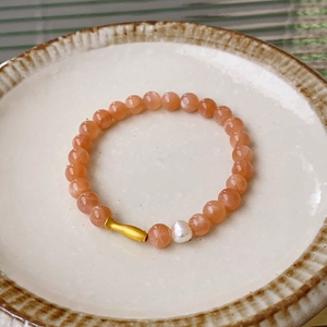 Detail of beaded bracelet featuring orange ore stones, a white pearl, and a gold fish charm