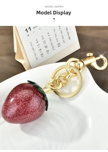 Detailed view of a sparkling red strawberry keychain with gold hardware on a ceramic tray
