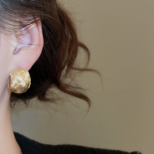 Close-up of golden C-shaped stud earrings with intricate woven texture worn on ear