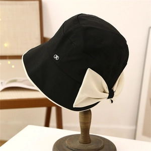 Detail of elegant black bucket hat with large cream bow and silver logo on wooden stand