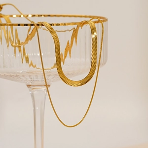 Detail of layered gold snake chains draped over a glass rim on a beige background