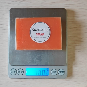 Digital scale showing Kojic Acid Soap net weight of 100g on a wood surface