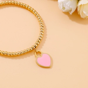 Detail of 18k gold plated beaded bracelet with a pink enamel heart charm on pink background