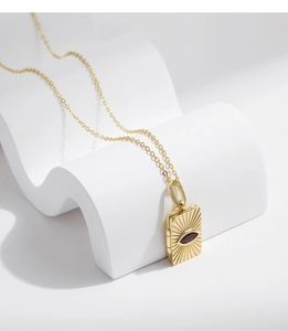 Macro detail of a gold sunburst envelope pendant with a deep red gemstone on a white wave