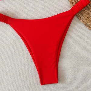 Détail du bas de bikini rouge échancré avec côtés froncés sur un fond de plage texturé