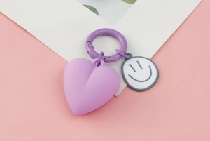 Close-up of a purple heart keychain with a grey smiley face pendant on a pink surface