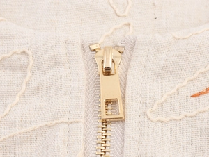 Detailed close-up of golden metal zipper on beige embroidered cotton linen fabric