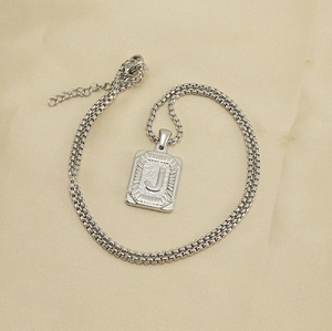 Detailed shot of silver square pendant with letter J and box chain on beige fabric