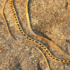 Drei geschichtete 18k Gold-Edelstahlketten auf einem natürlichen braunen Steinboden