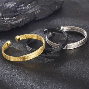 Stack of gold, silver, and black stainless steel cable open bangles on a stone texture