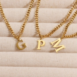 Detail of 18K gold plated serif letter pendants G, P, and M on a beige jewelry display