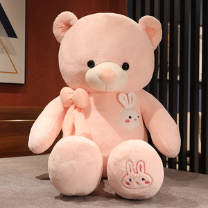 Detailed view of pink plush teddy bear with white rabbit patch and bow on a cozy bed