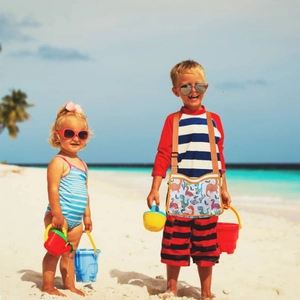 Kids dinosaur mesh beach bag with orange strap shown in a sunny outdoor seaside setting